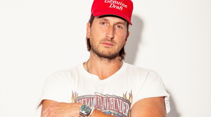 A man in a white graphic tshirt and red cap stood in front of a white wall